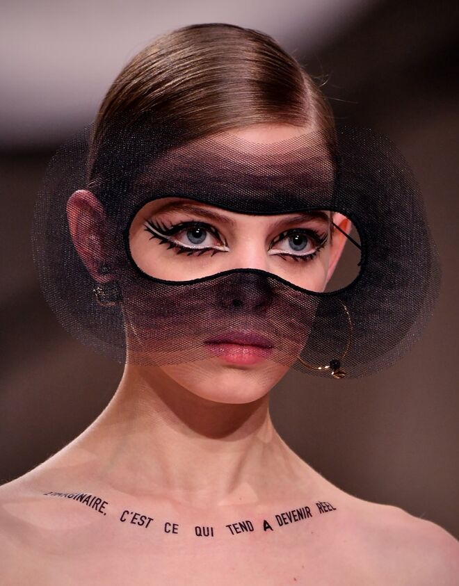 Desfile alta-costura primavera/verão 2018 Dior: Os cabelos foram perfeitamente penteados e presos em coque, com uma risca lateral. As modelos tinham frases inspiradoras escritas pelo corpo.