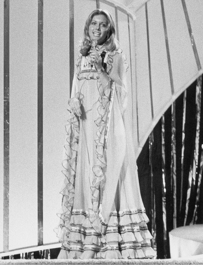 Aquela que anos depois foi estrela mundial em “Grease” participou na Eurovisão e o visual era também muito anos 70. Usou um vestido azul que bem podia ser uma camisa de noite, cheio de folhos. Ficou em quarto lugar com a sua música "Long Live Love".