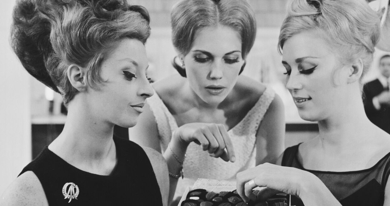 Três mulheres observam uma caixa de bombons, com penteados elaborados