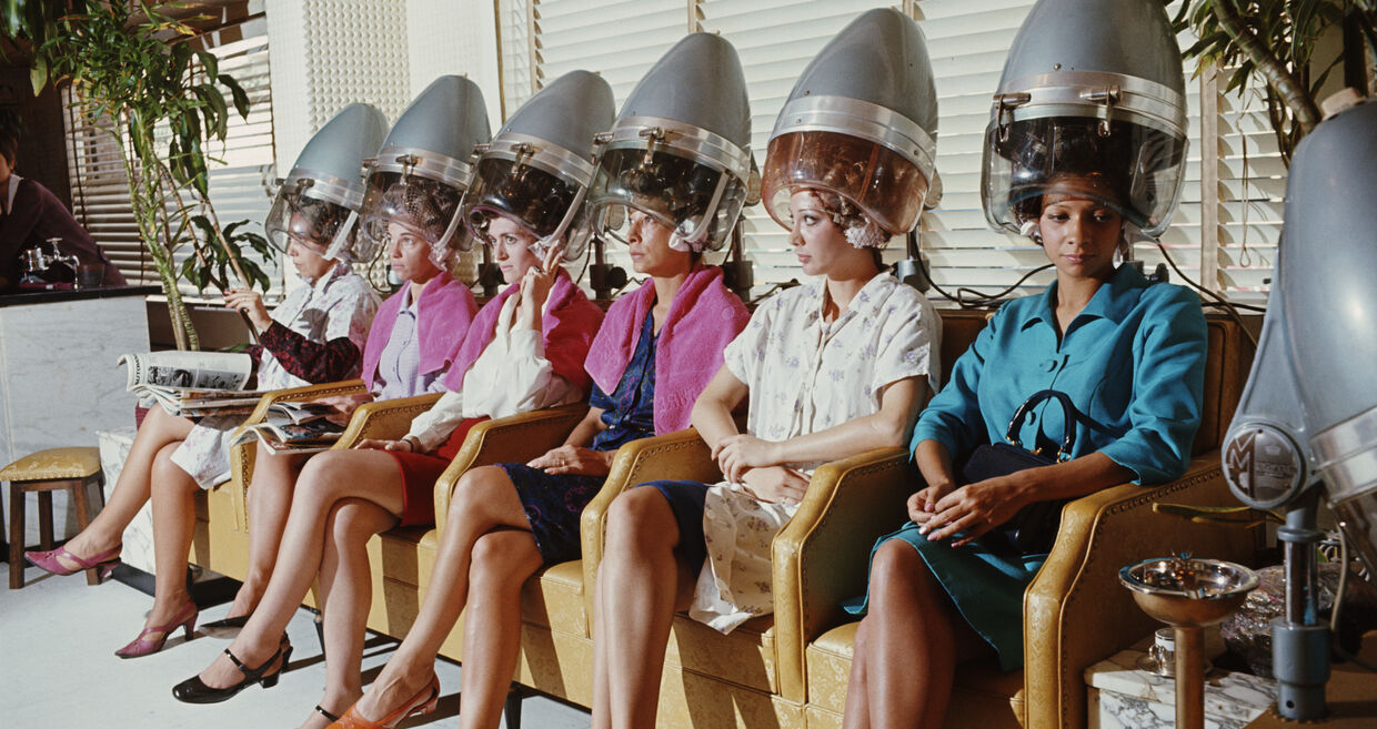 Num salão de beleza, mulheres aguardam com secadores de cabelo