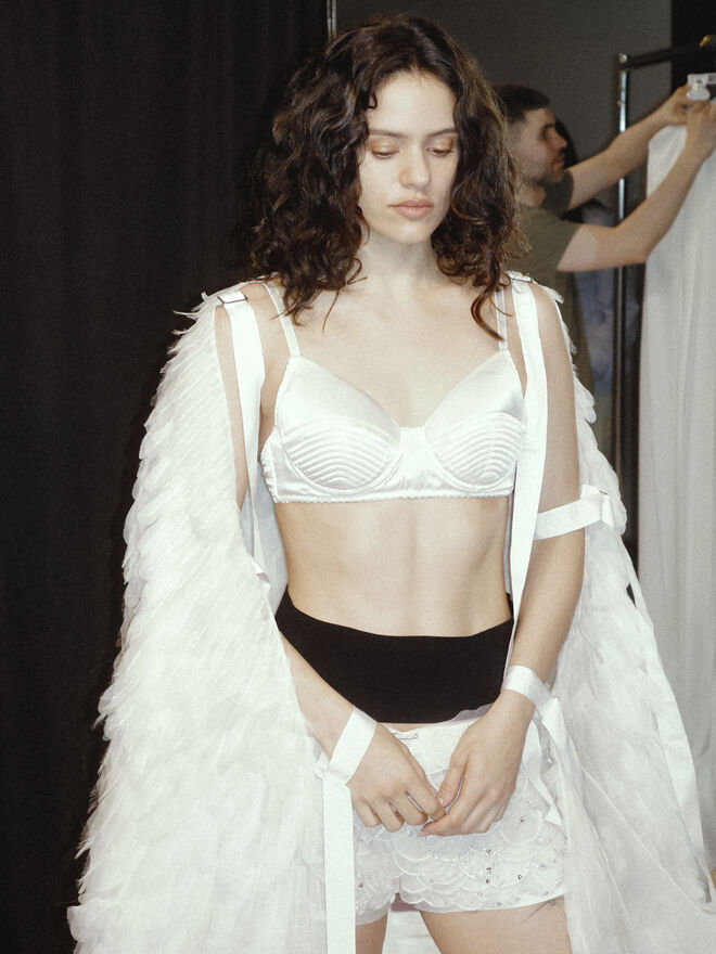  Rosalía veste uma capa inspirada em asas de anjo, construída em organza e penas de chiffon, combinada com um soutien branco em cetim de acabamento acolchoado e uns calções bordados com escamas de organza marfim, enriquecidas com lantejoulas e missangas prateadas